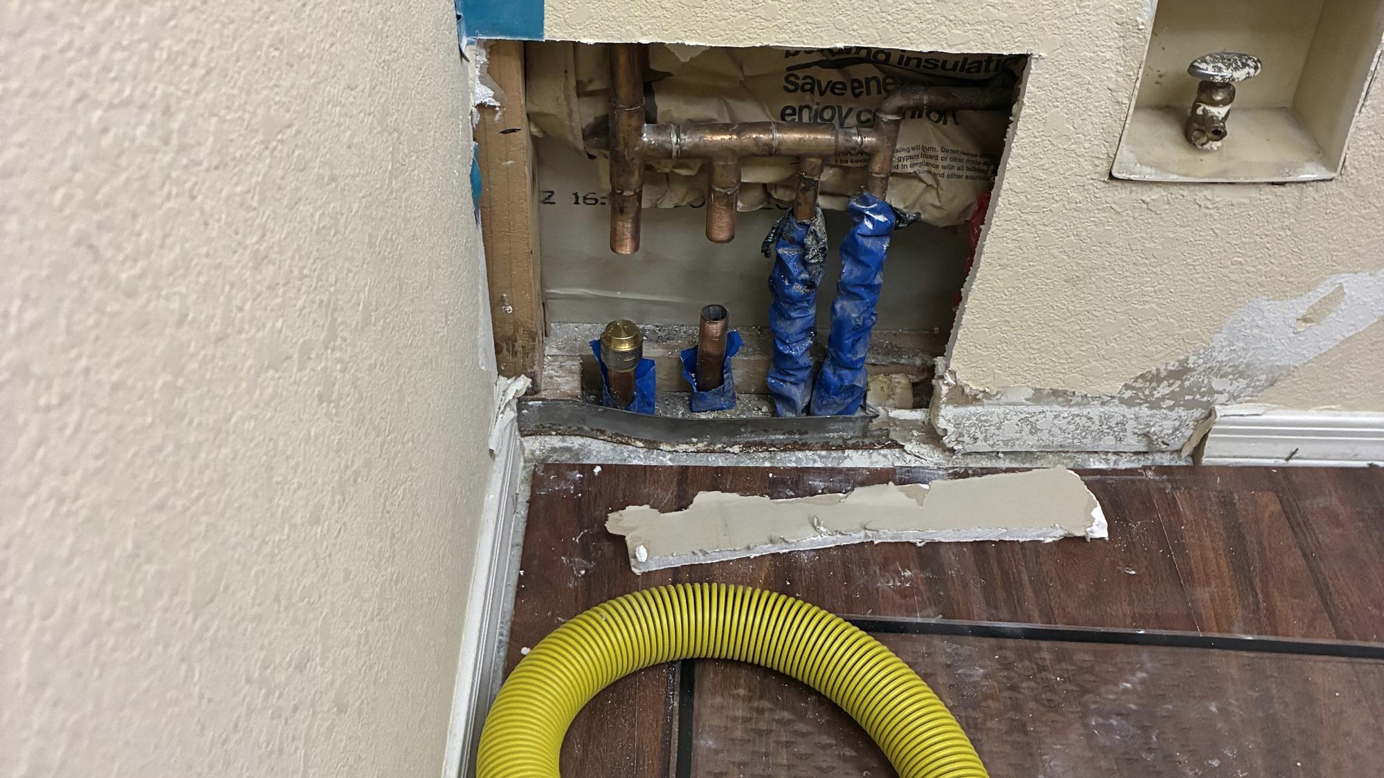 An opened interior wall with copper supply lines and a drying hose run into the cavity
