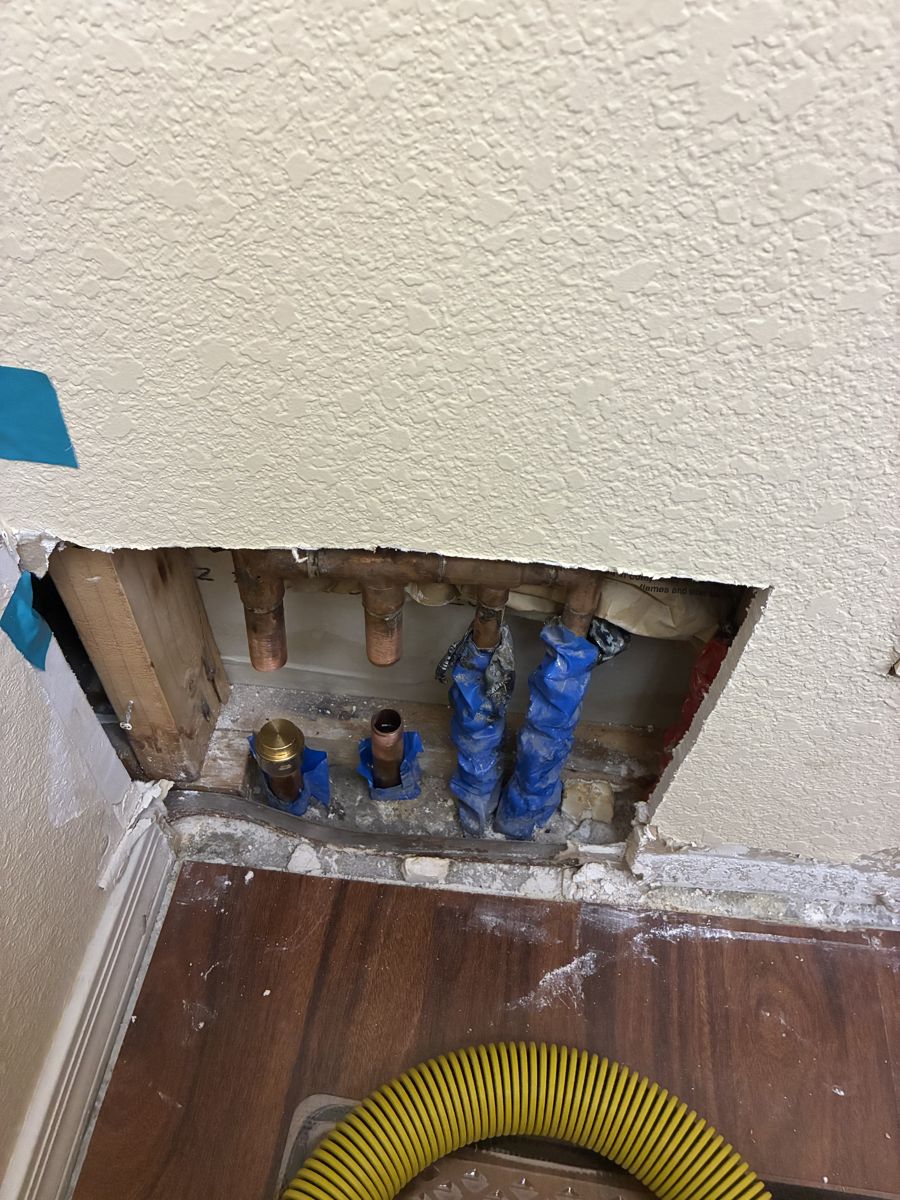 Opened interior wall showing copper supply stubs wrapped in blue tape with a drying hose run into the cavity